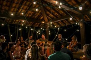 Público reunido em roda musical ao ar livre sob varal de lâmpadas enquanto um músico toca violão