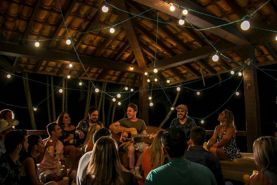 Público reunido em roda musical ao ar livre sob varal de lâmpadas enquanto um músico toca violão