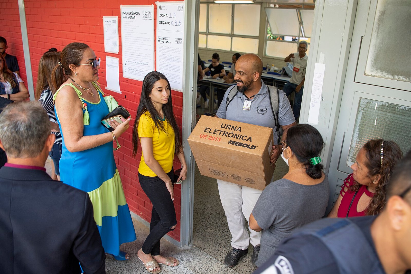 Mesário carrega caixa de urna eletrônica na entrada de uma seção eleitoral, ao lado de eleitores na fila