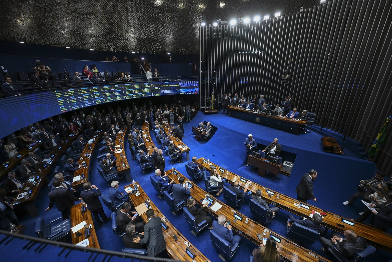 Vista do alto do plenário do Senado Federal em sessão, com senadores nas bancadas de madeira, tapete azul e painel eletrônico de votação ao fundo