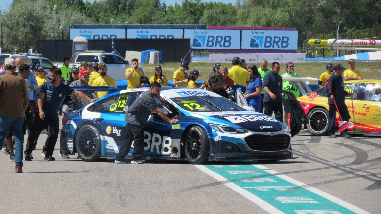 Mecânico empurra o Chevrolet Cruze azul número 12 da Stock Car, com patrocínio do BRB, no pitlane, cercado por equipes de uniforme amarelo