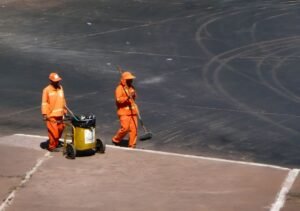 Dois garis de uniforme laranja trabalham em via urbana, um empurrando carrinho de coleta e outro varrendo o asfalto