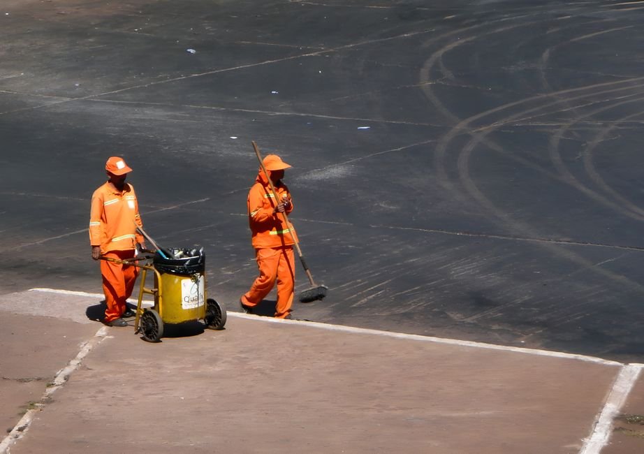 Dois garis de uniforme laranja trabalham em via urbana, um empurrando carrinho de coleta e outro varrendo o asfalto