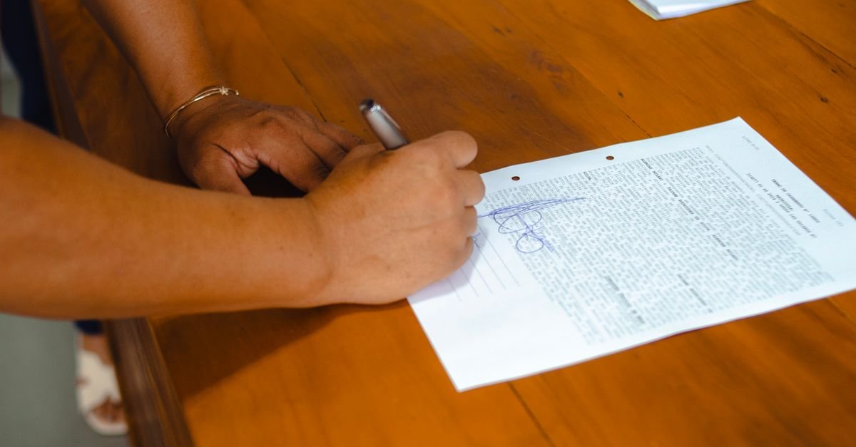 Mãos assinando um contrato impresso sobre uma mesa de madeira