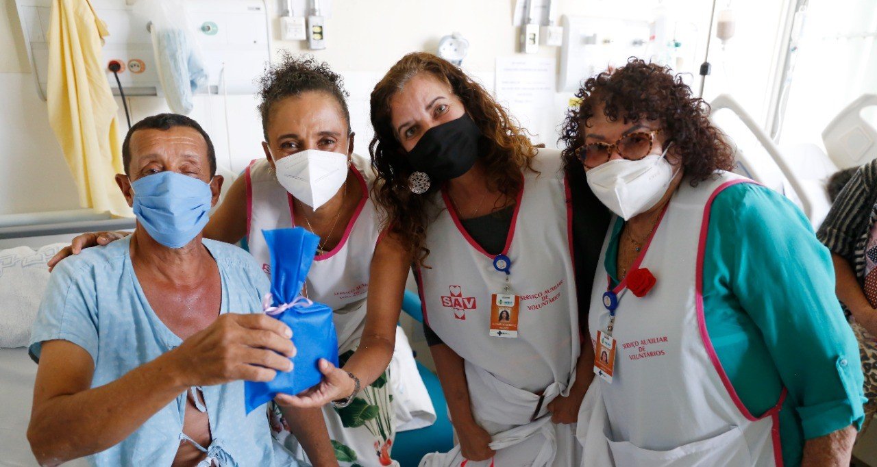 Três voluntárias do SAV com colete branco ao lado de paciente internado no Hospital de Base, que segura um kit de higiene