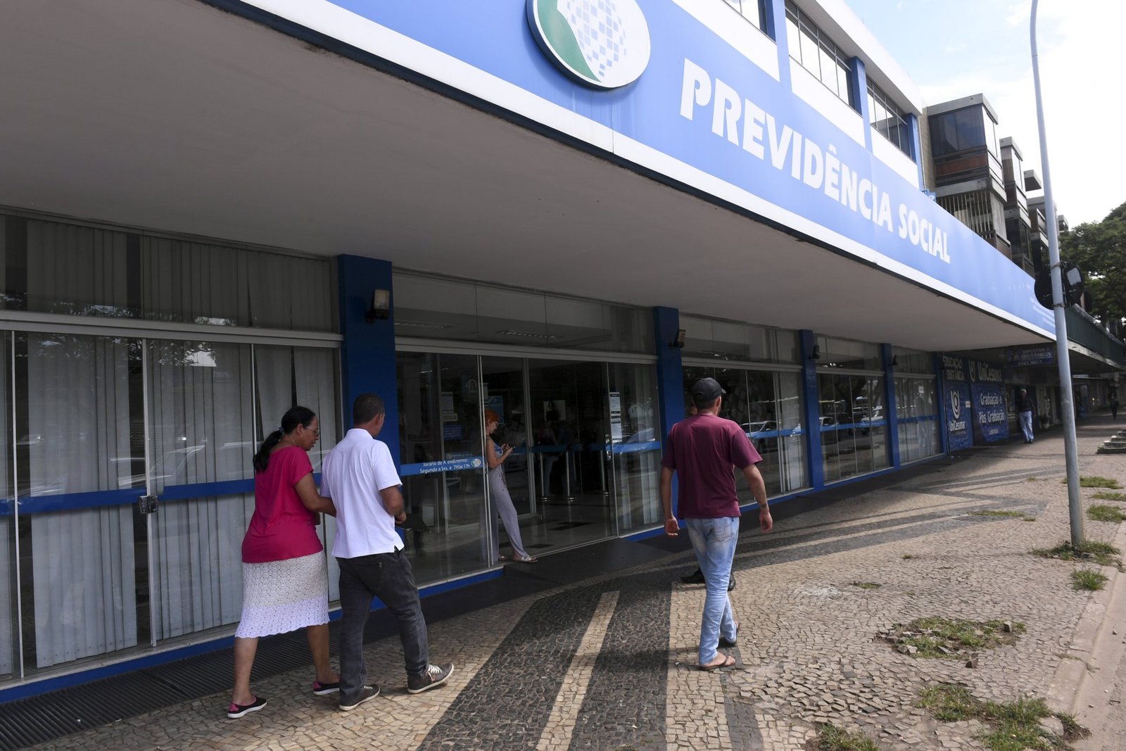 Pessoas caminham diante de uma agência da Previdência Social em Brasília, com a fachada azul e a porta de entrada ao fundo