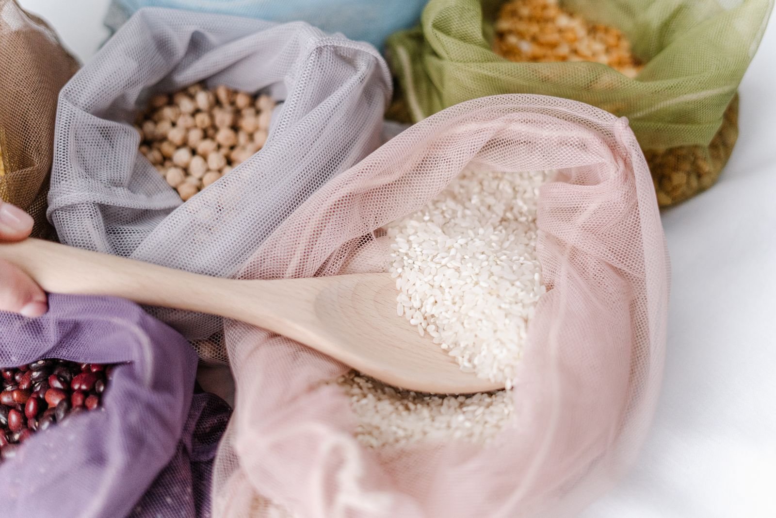 Colher de madeira em sacola de tela com arroz, ao lado de sacolas com feijão e grão-de-bico
