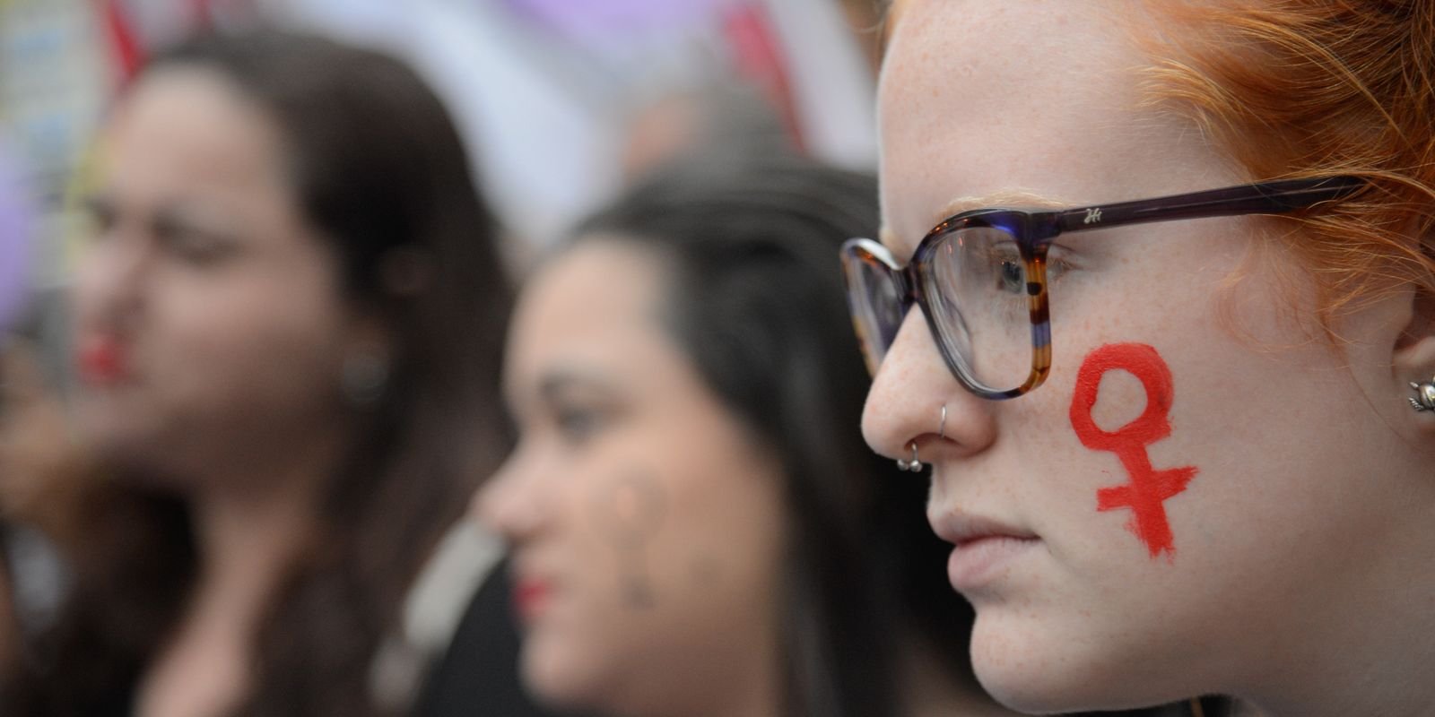 Mulheres em ato com o símbolo feminino pintado no rosto; foto de arquivo da Agência Brasil