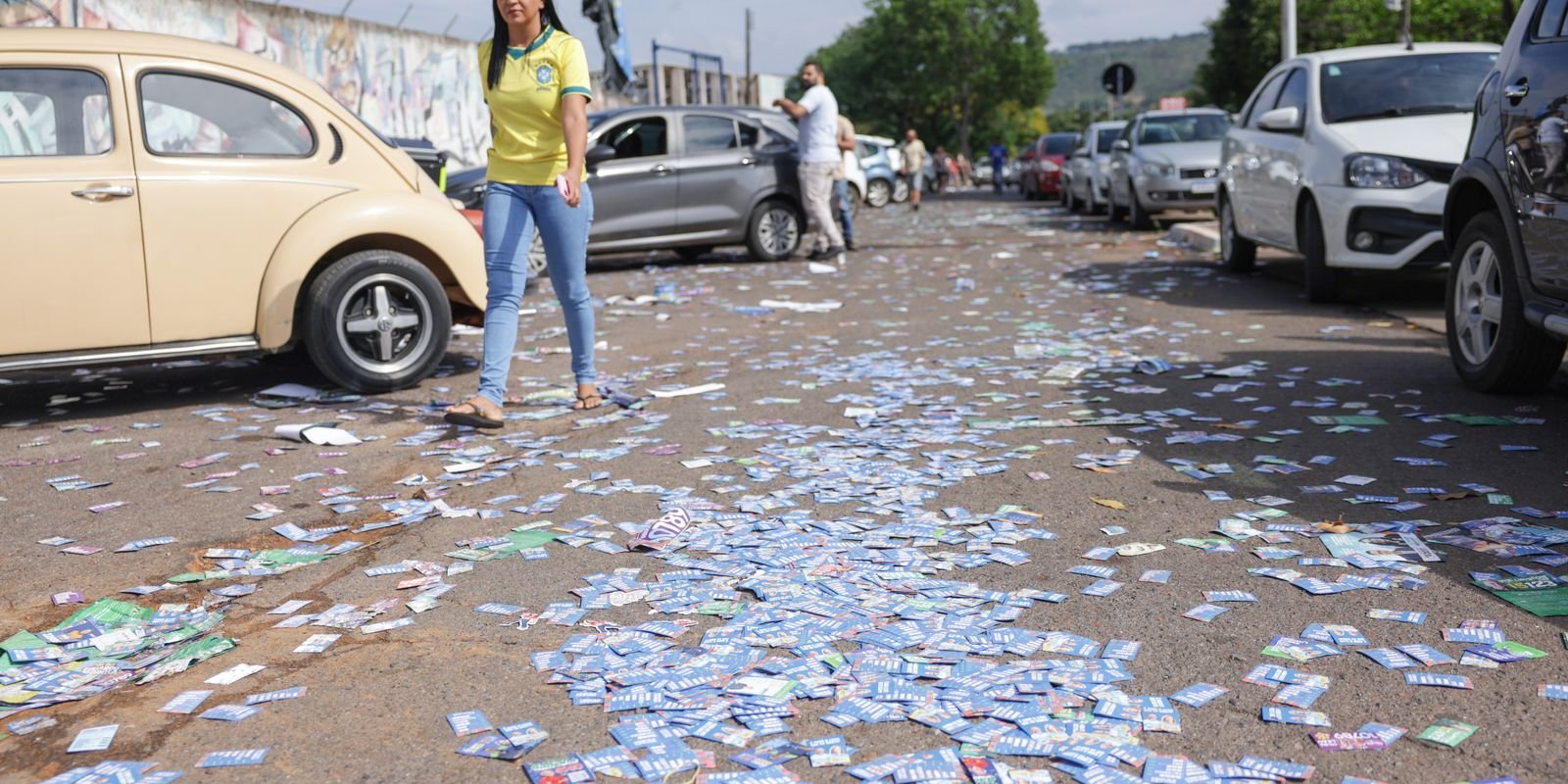 Santinhos de campanha espalhados no chão de uma rua durante o primeiro turno das eleições, com pedestres e carros estacionados