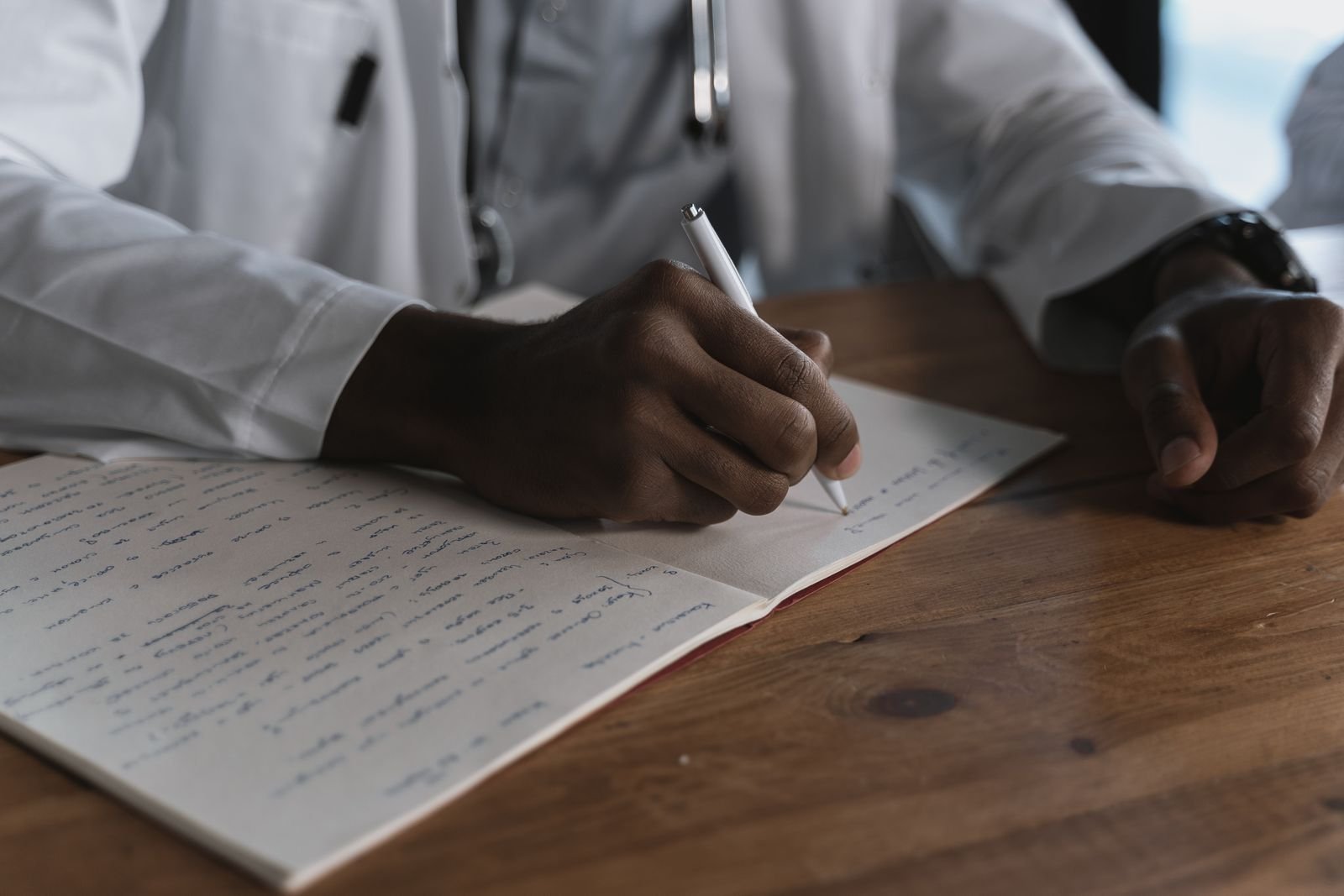 Mãos de médico escrevendo em caderno sobre mesa de madeira, foto ilustrativa