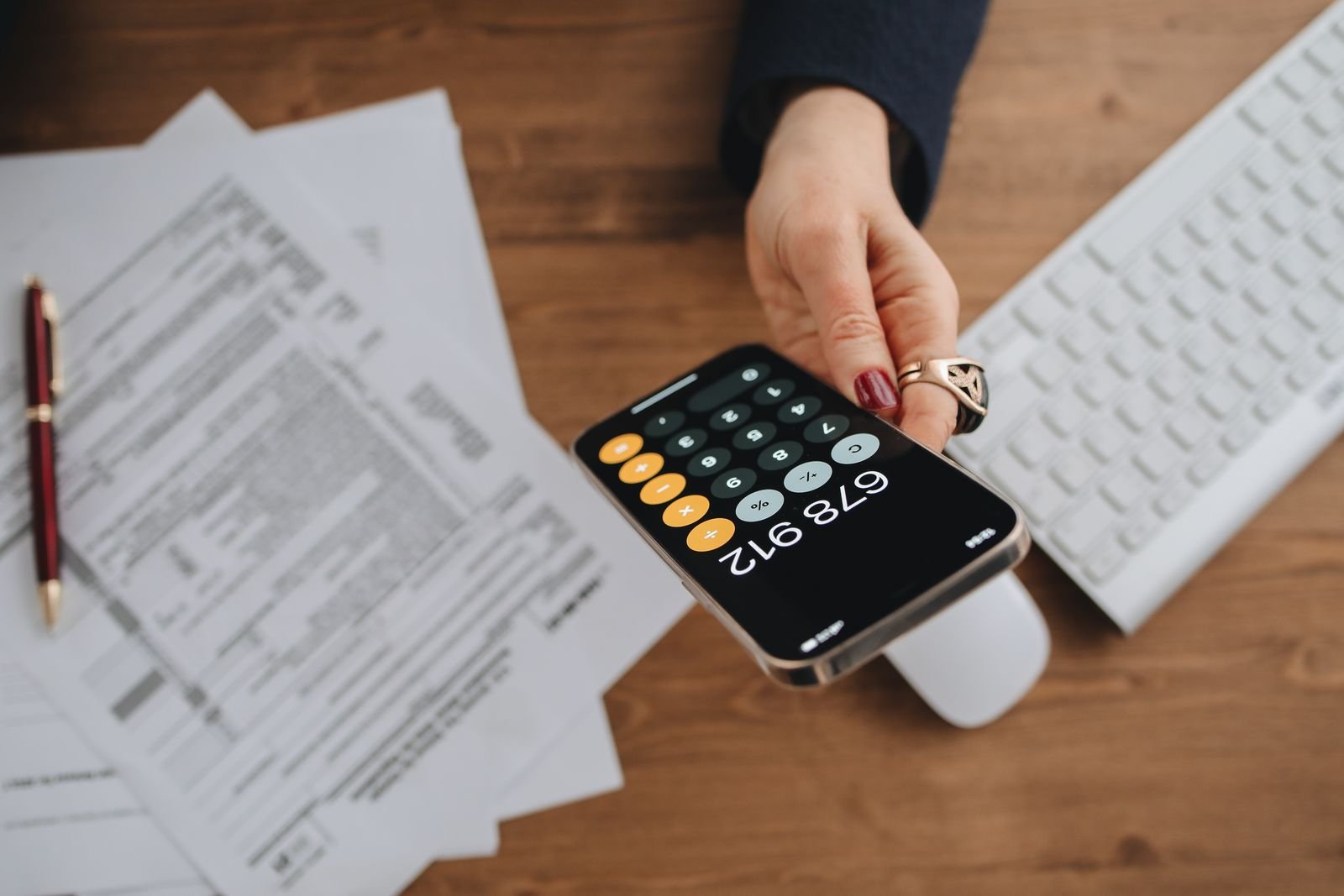 Mão segura um celular com a calculadora aberta sobre documentos fiscais em uma mesa de madeira
