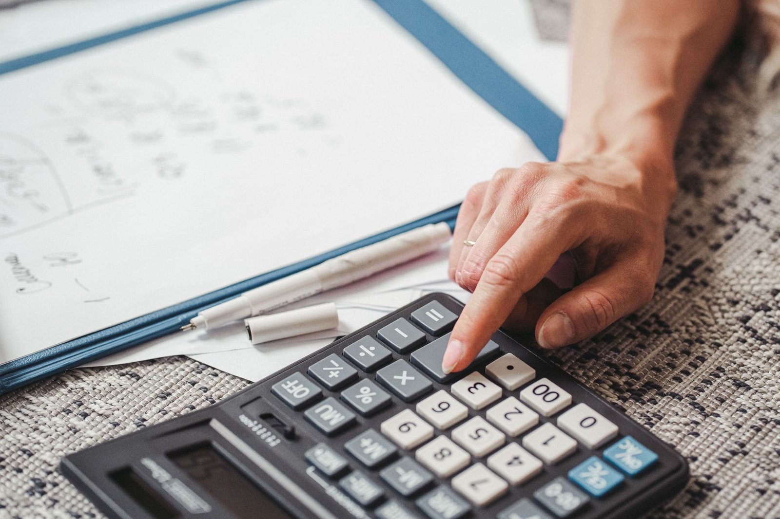 Mão digitando em uma calculadora sobre papéis e uma pasta azul