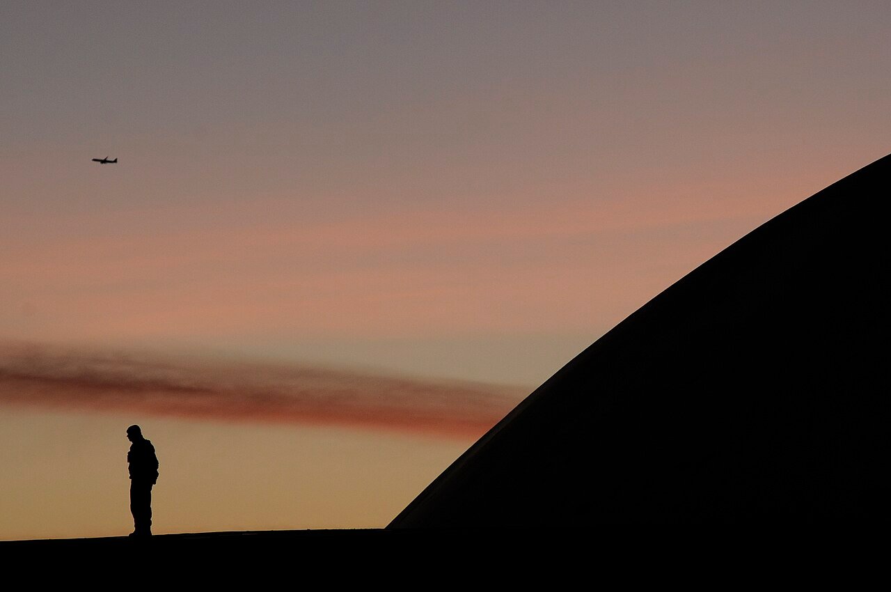 Silhueta de uma pessoa ao lado da cúpula do Senado, no Congresso Nacional, em Brasília, ao entardecer, com um avião no céu; foto de arquivo