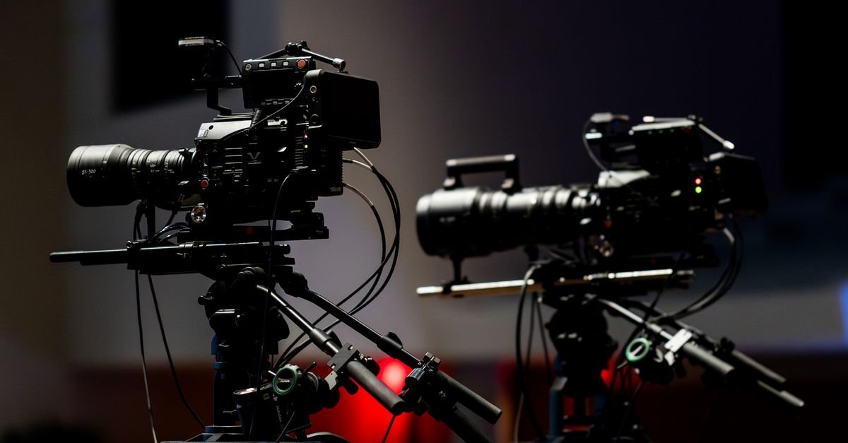 Duas câmeras profissionais de vídeo em tripés, em estúdio de televisão com iluminação baixa (foto ilustrativa)