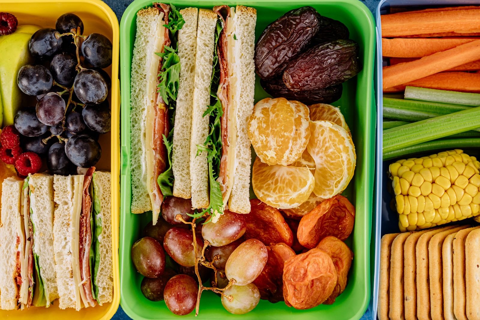 Marmitas com sanduíches, uvas, tangerina, cenoura e milho, exemplo de lanche escolar
