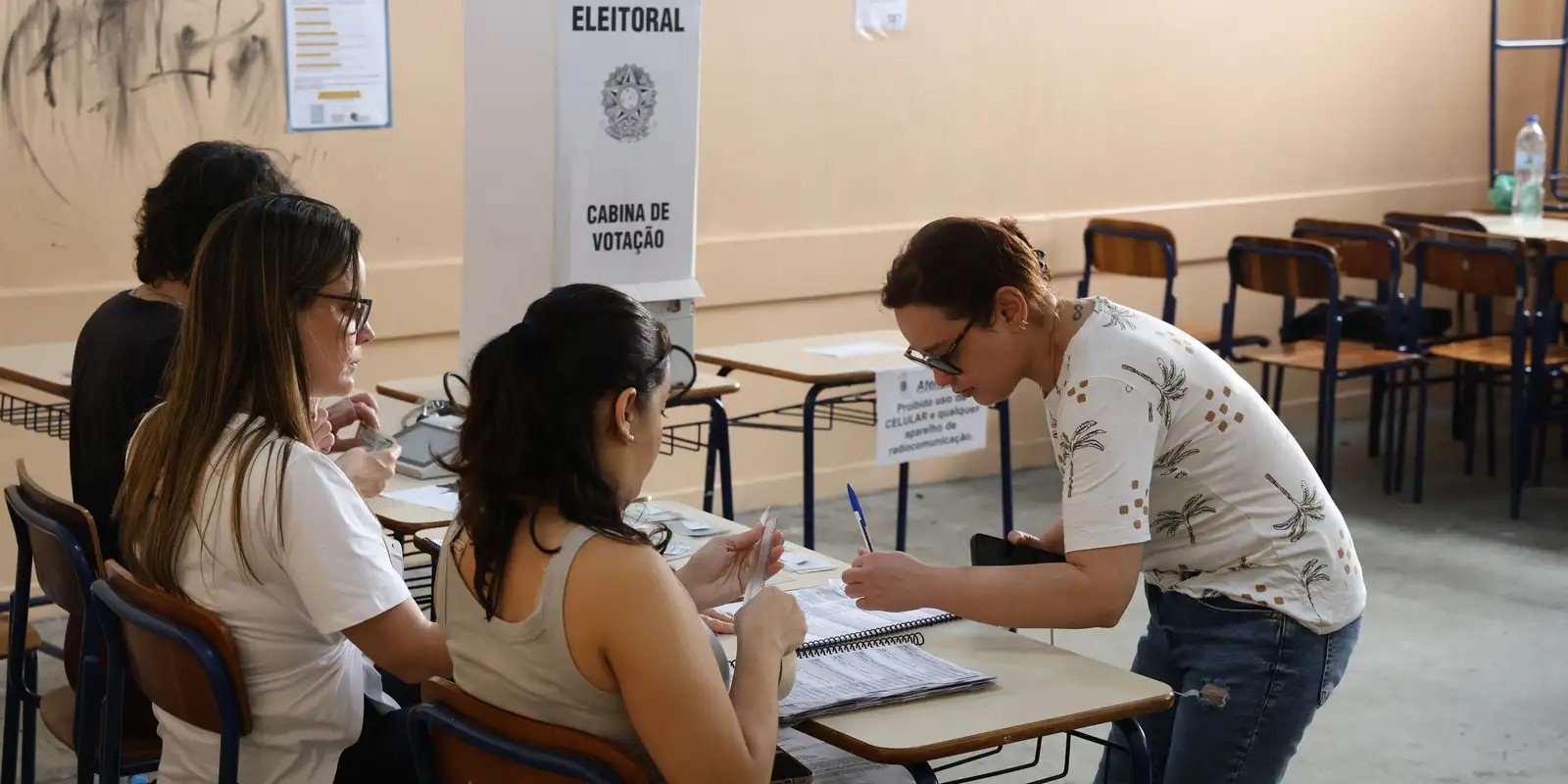 Eleitor assina o caderno de votação diante de duas mesárias, ao lado de uma cabine de votação, durante o primeiro turno de 2026