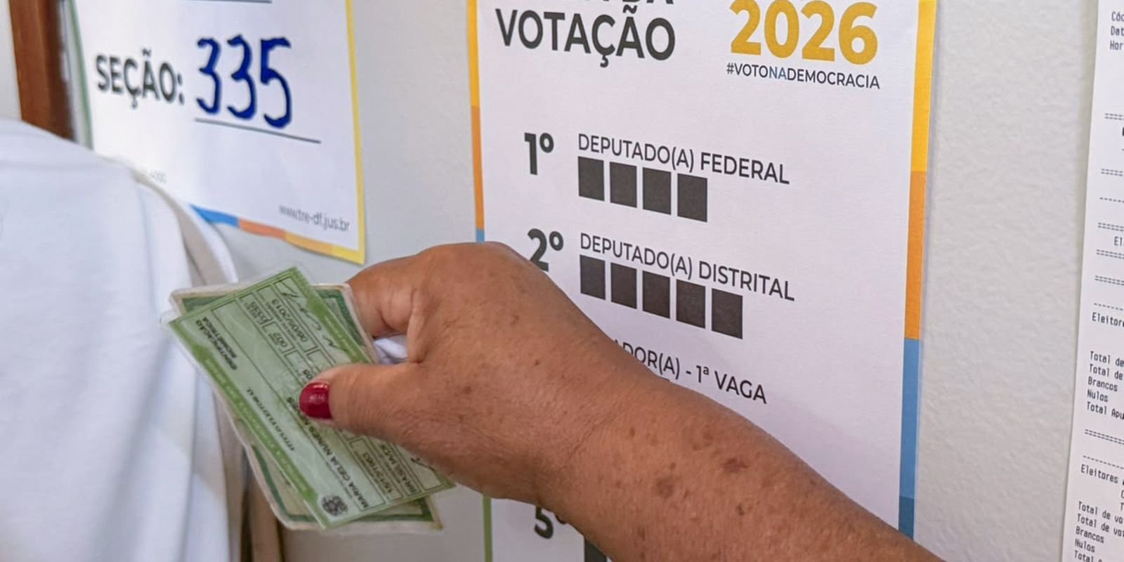 Eleitora segura o título de eleitor diante do cartaz com a ordem de votação de 2026 em uma seção eleitoral do DF
