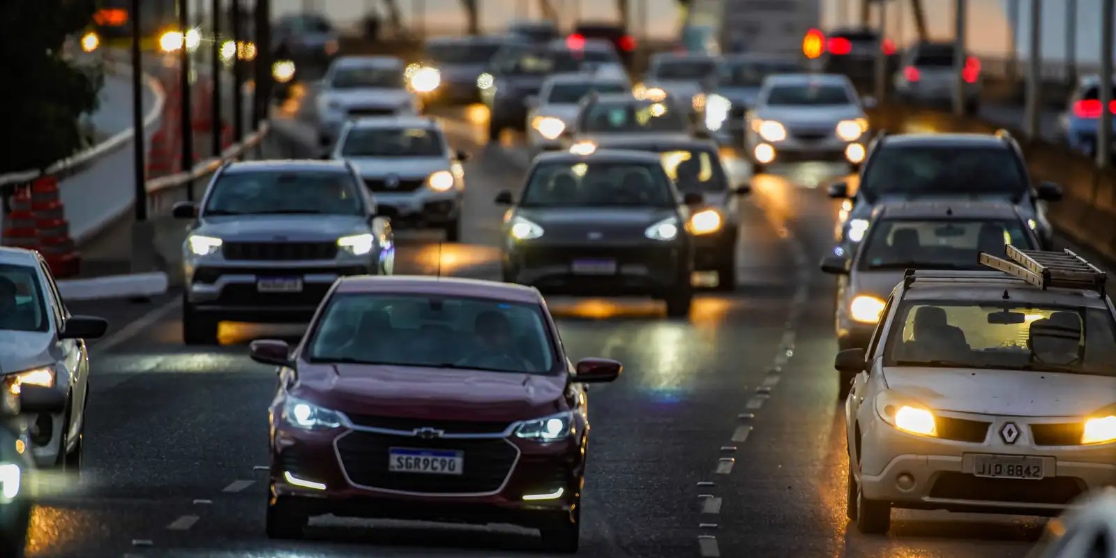 Carros em fila de trânsito ao fim da tarde, com faróis acesos