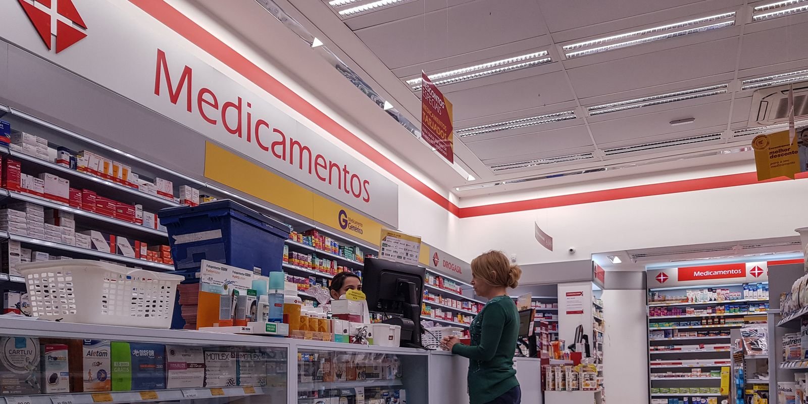 Atendente de farmácia conversa com cliente em frente à prateleira de medicamentos