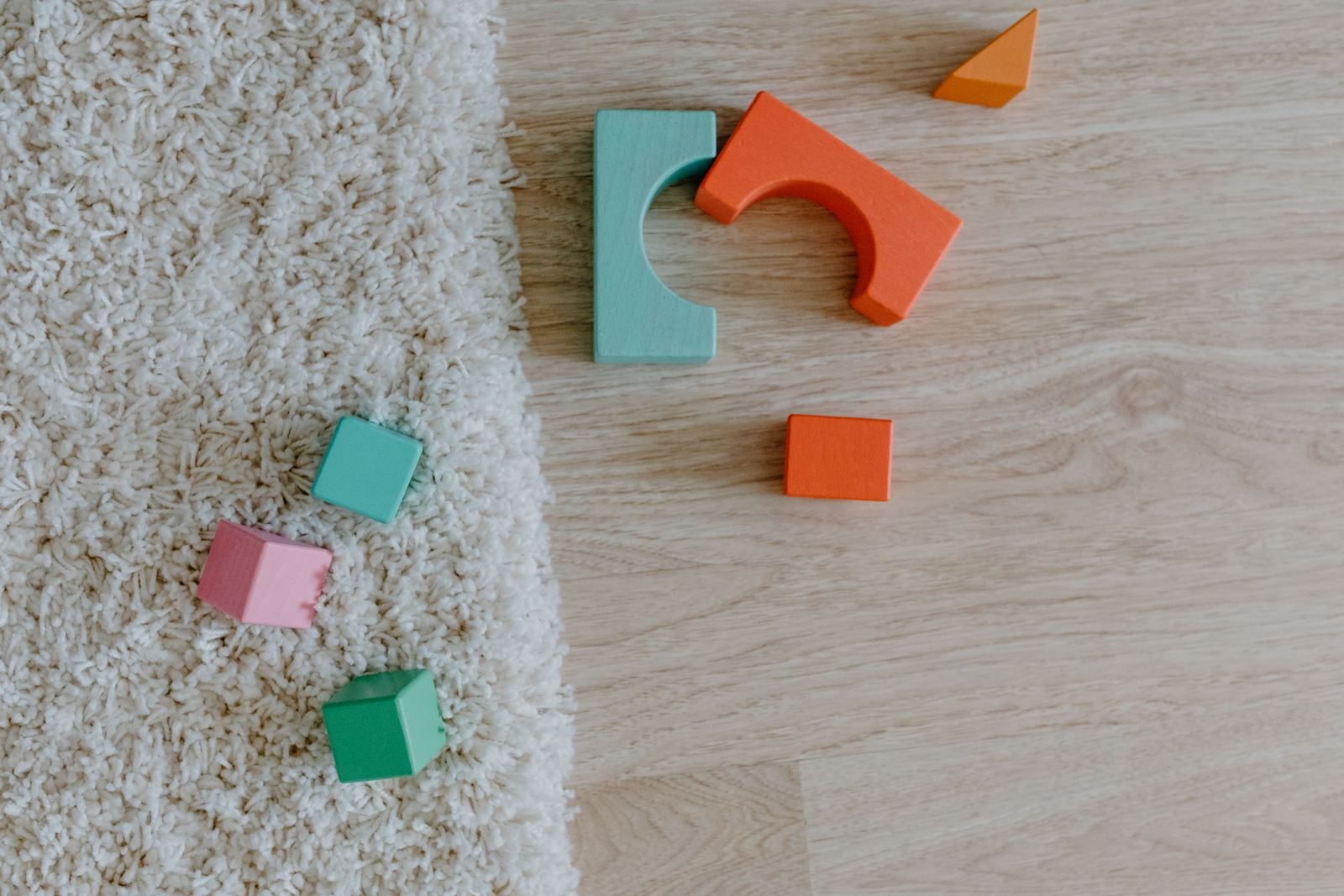 Blocos de madeira coloridos sobre tapete e piso, em imagem ilustrativa de sala de creche