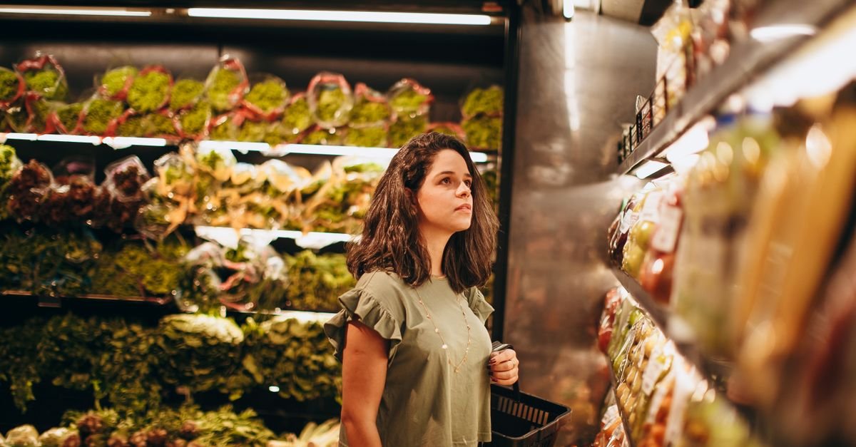 Mulher com cesta de compras observa verduras e legumes na seção de hortifrúti de um supermercado, em imagem ilustrativa