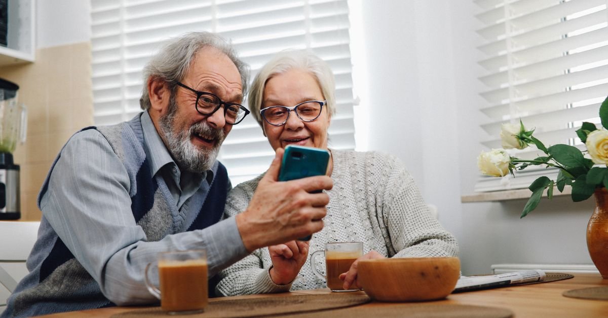 Casal de idosos sorrindo olha para um celular sobre a mesa da cozinha (foto ilustrativa)