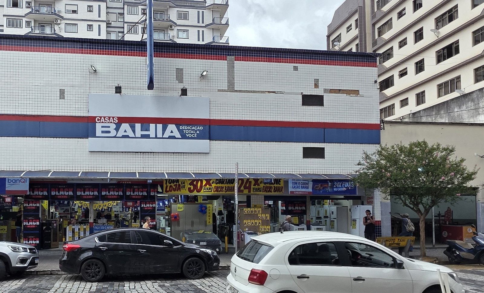 Fachada de uma loja das Casas Bahia, com placa da rede e faixas de promoção, em rua de paralelepípedo