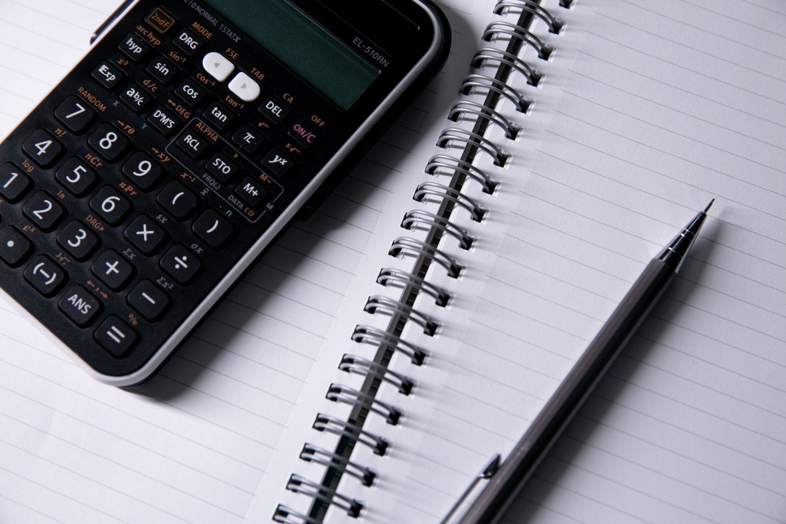Calculadora ao lado de um caderno de espiral aberto e uma caneta sobre uma mesa