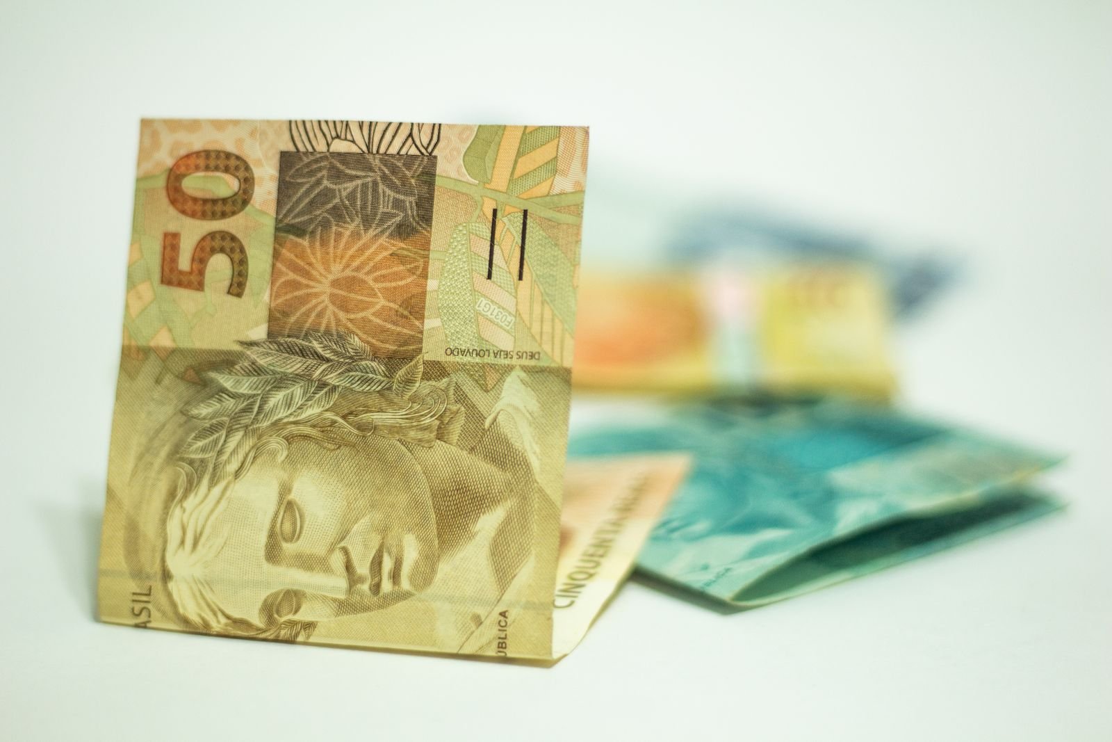 Cédula de R$ 50 em pé sobre superfície branca, com outras notas de real desfocadas ao fundo