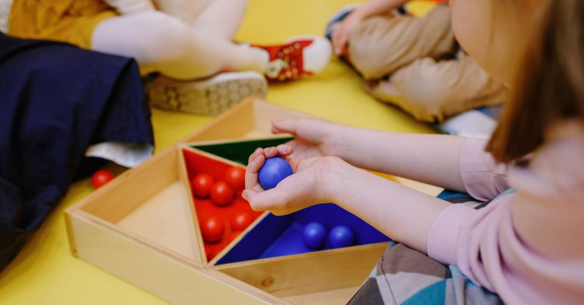 Mãos de adulto mostram brinquedos de madeira educativos a bebês sentados em um tapete amarelo; imagem ilustrativa