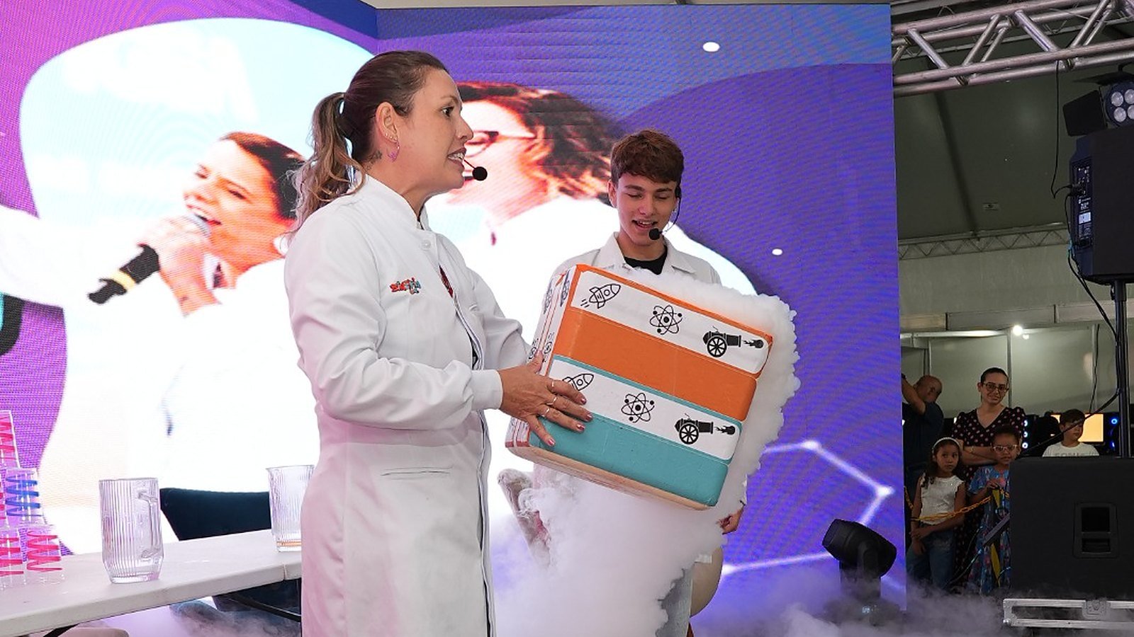 Apresentadora de jaleco segura caixa colorida que solta fumaça durante experimento no palco do Ciência na Estrada