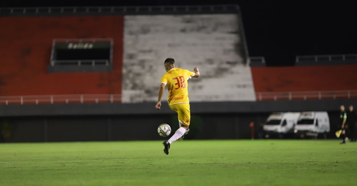 Jogador de camisa amarela chuta a bola em campo de futebol iluminado, à noite