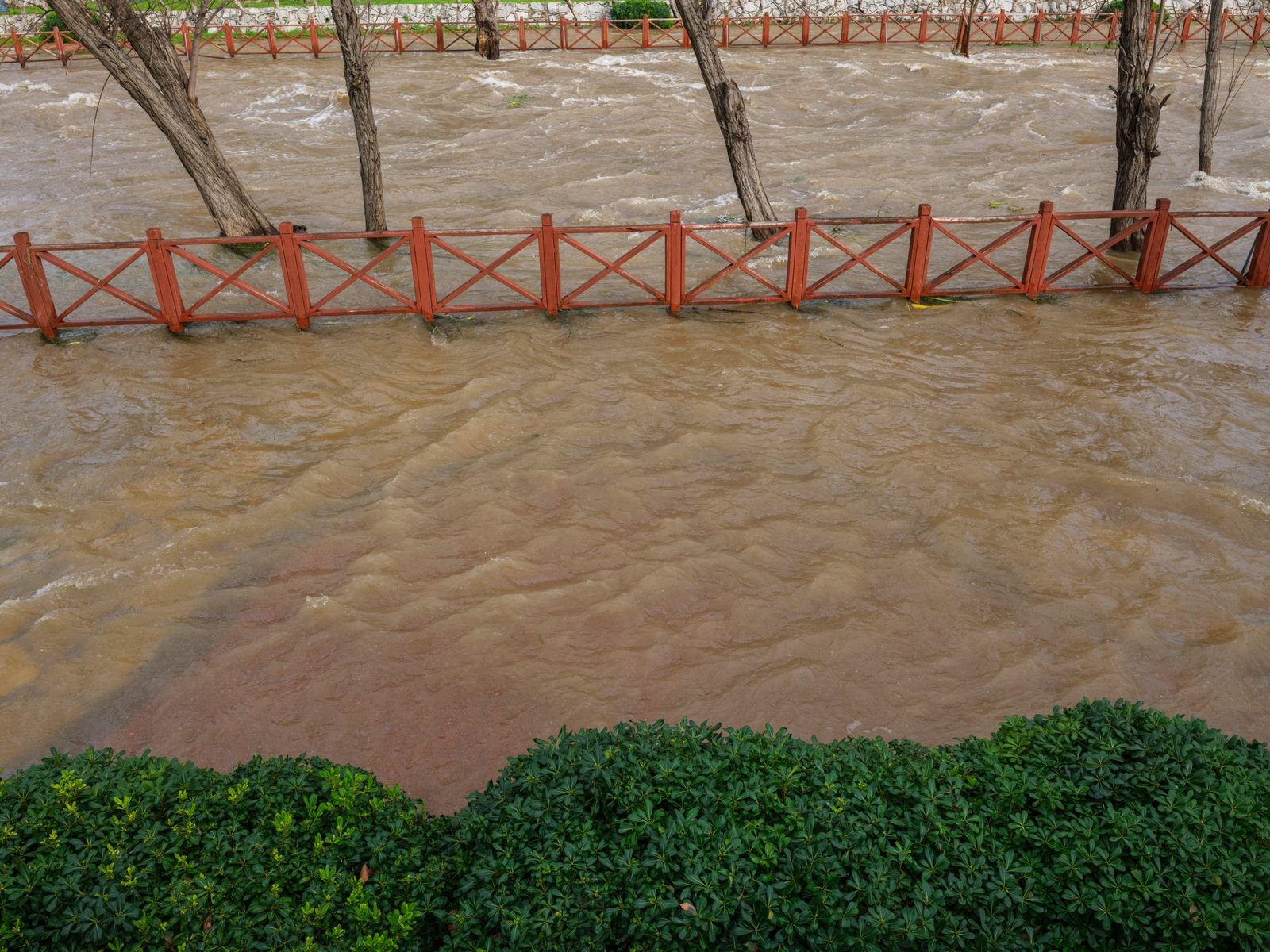 Córrego de águas barrentas transbordando junto a cercas vermelhas e árvores em um parque; foto ilustrativa, não é do local