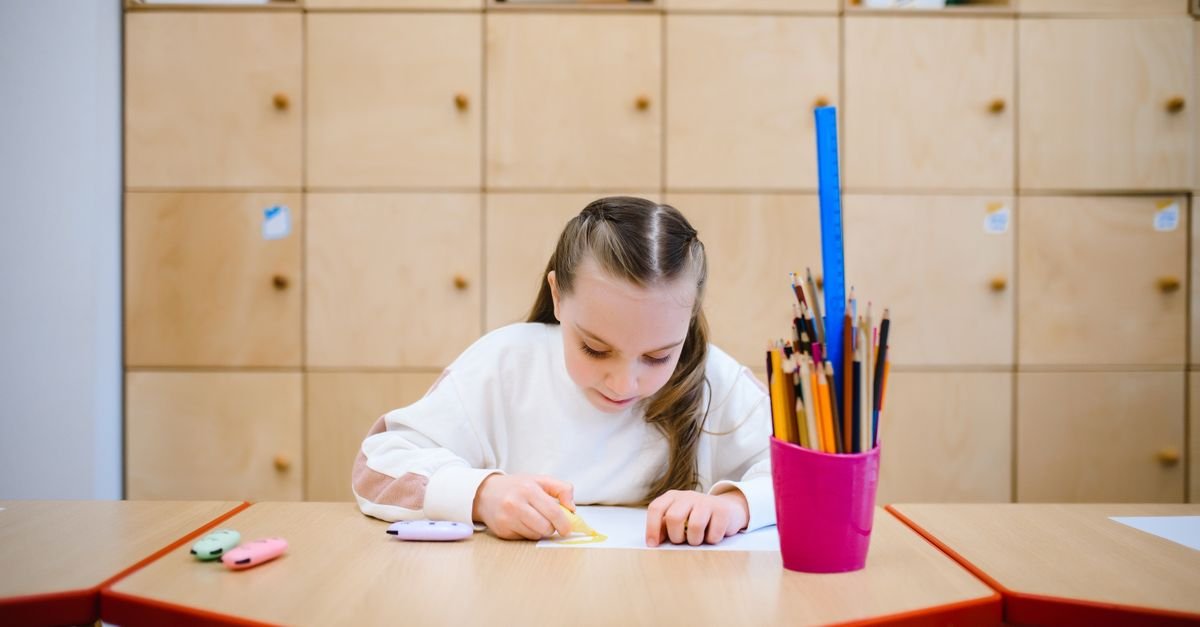 Menina desenha com lápis de cor em uma mesa de sala de aula, com armário de madeira ao fundo