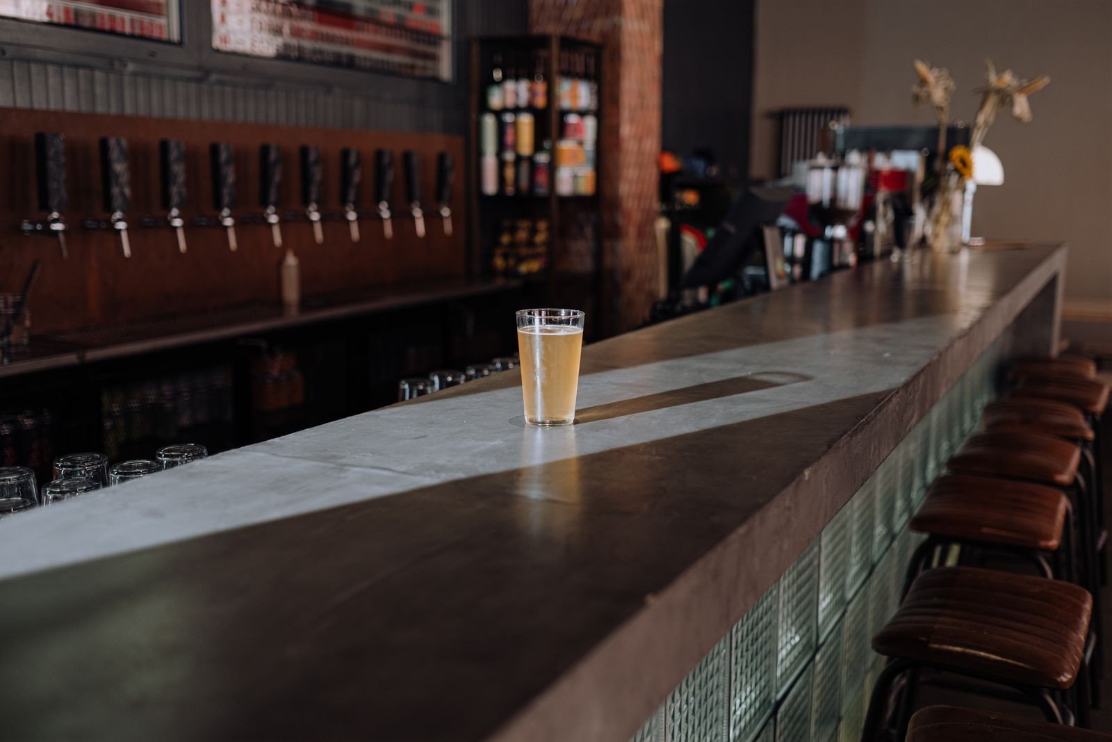 Copo de cerveja sobre o balcão de um bar vazio, com torneiras de chope ao fundo, imagem ilustrativa