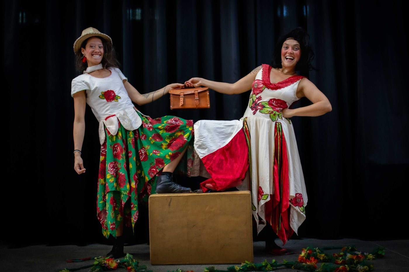 Duas artistas palhaças do grupo As Pareias, de saias floridas e com uma mala, em palco escuro