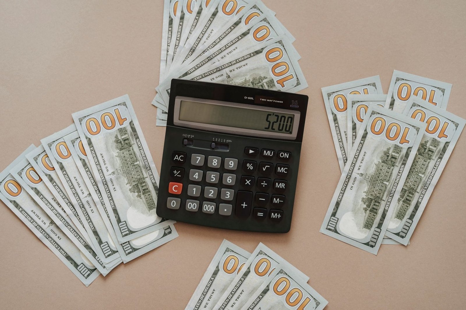 Calculadora com o visor em 5.200 cercada por notas de cem dólares sobre fundo bege
