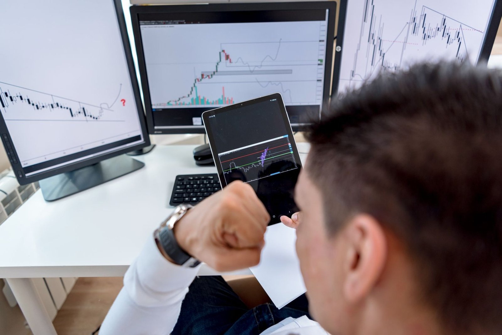 Homem observa gráficos de negociação de ações em monitores e em um tablet, imagem ilustrativa de mercado financeiro