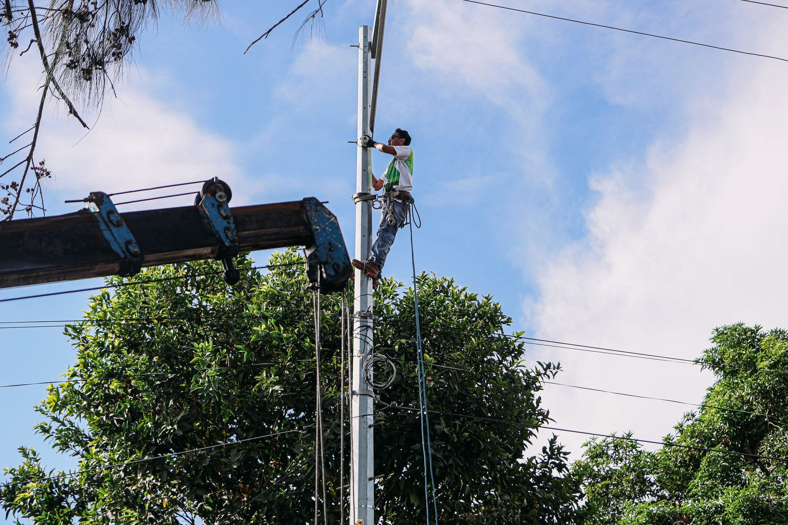 Eletricista em trabalho no alto de um poste de rede elétrica, com árvores e céu azul ao fundo