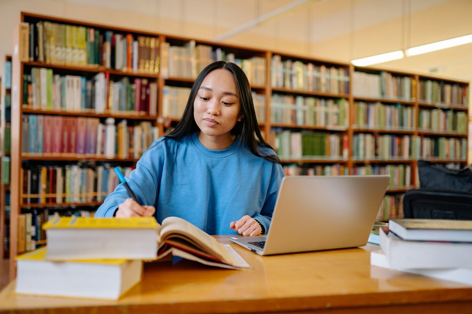 Estudante universitária escrevendo em um livro, com notebook, em uma biblioteca