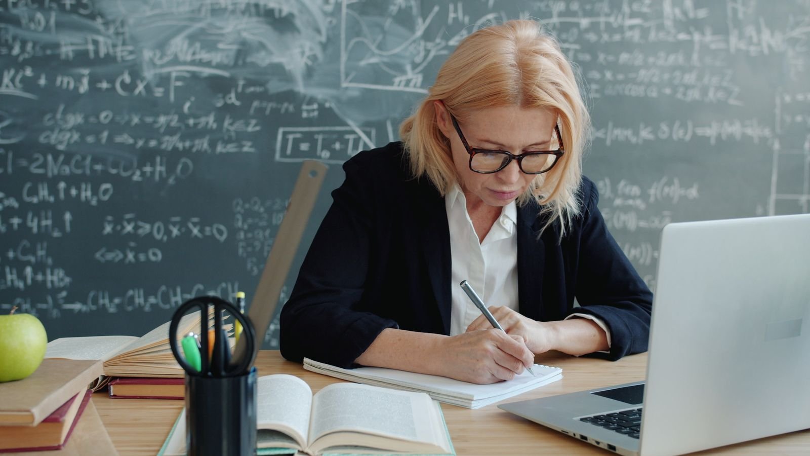 Professora escreve em caderno diante de lousa com fórmulas, com livros e notebook sobre a mesa; foto de arquivo
