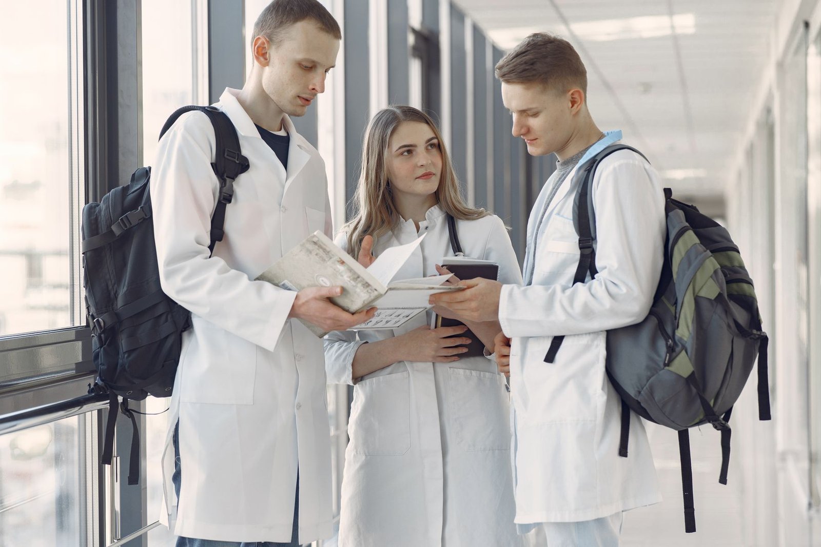 Três estudantes de medicina de jaleco conversam em um corredor, com mochilas e um livro aberto