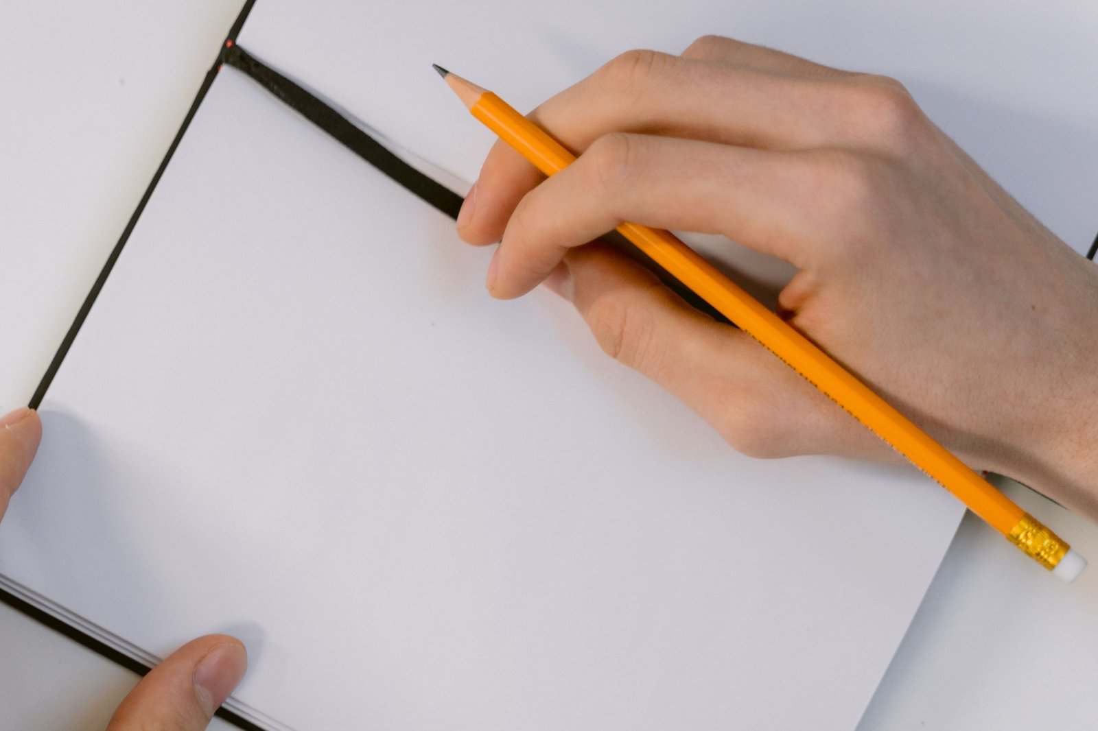 Mão segurando um lápis amarelo sobre um caderno aberto de páginas em branco, em uma mesa de estudo