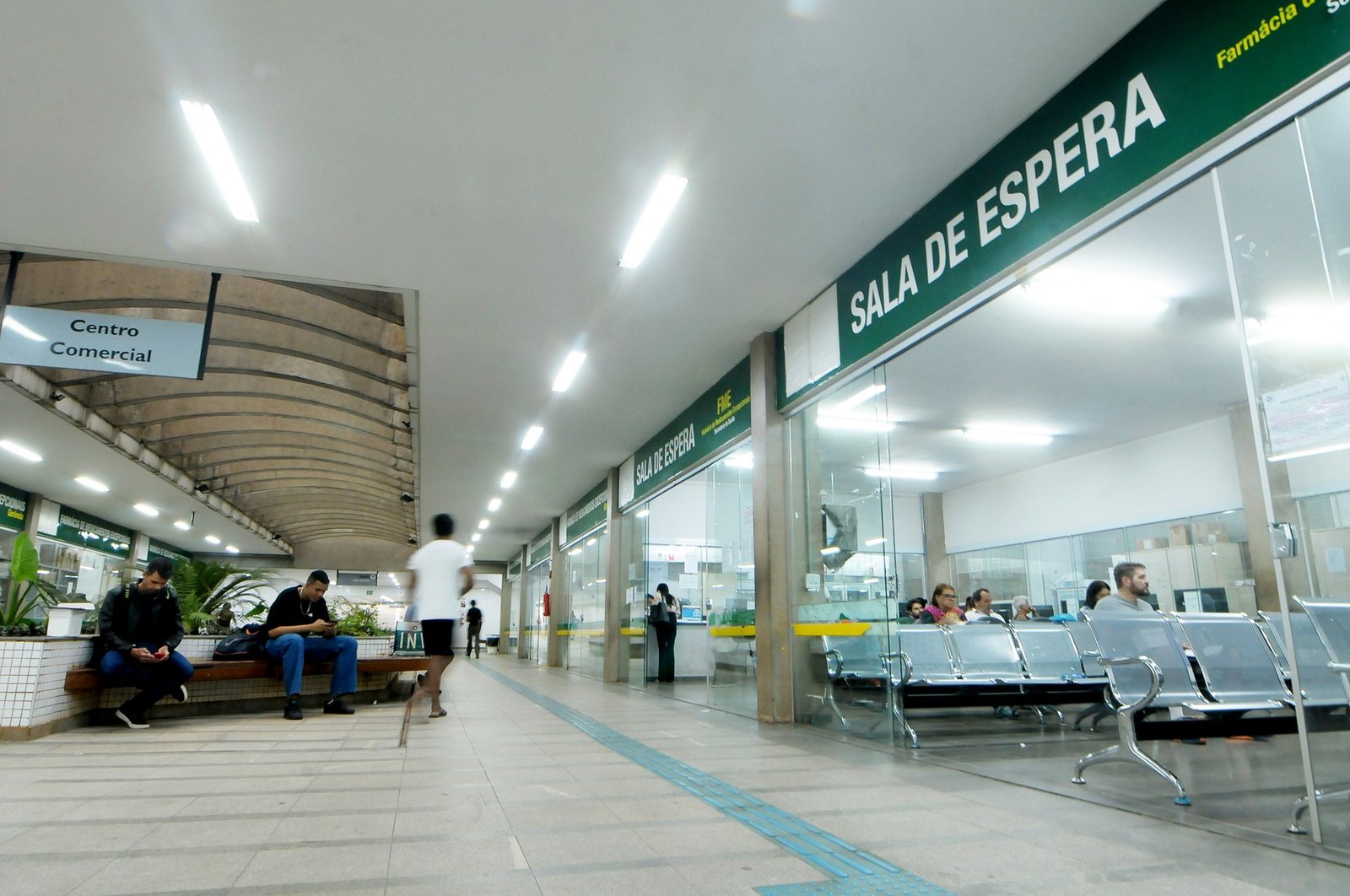 Sala de espera de uma Farmácia de Alto Custo do DF, com pacientes sentados e placas de sinalização verdes