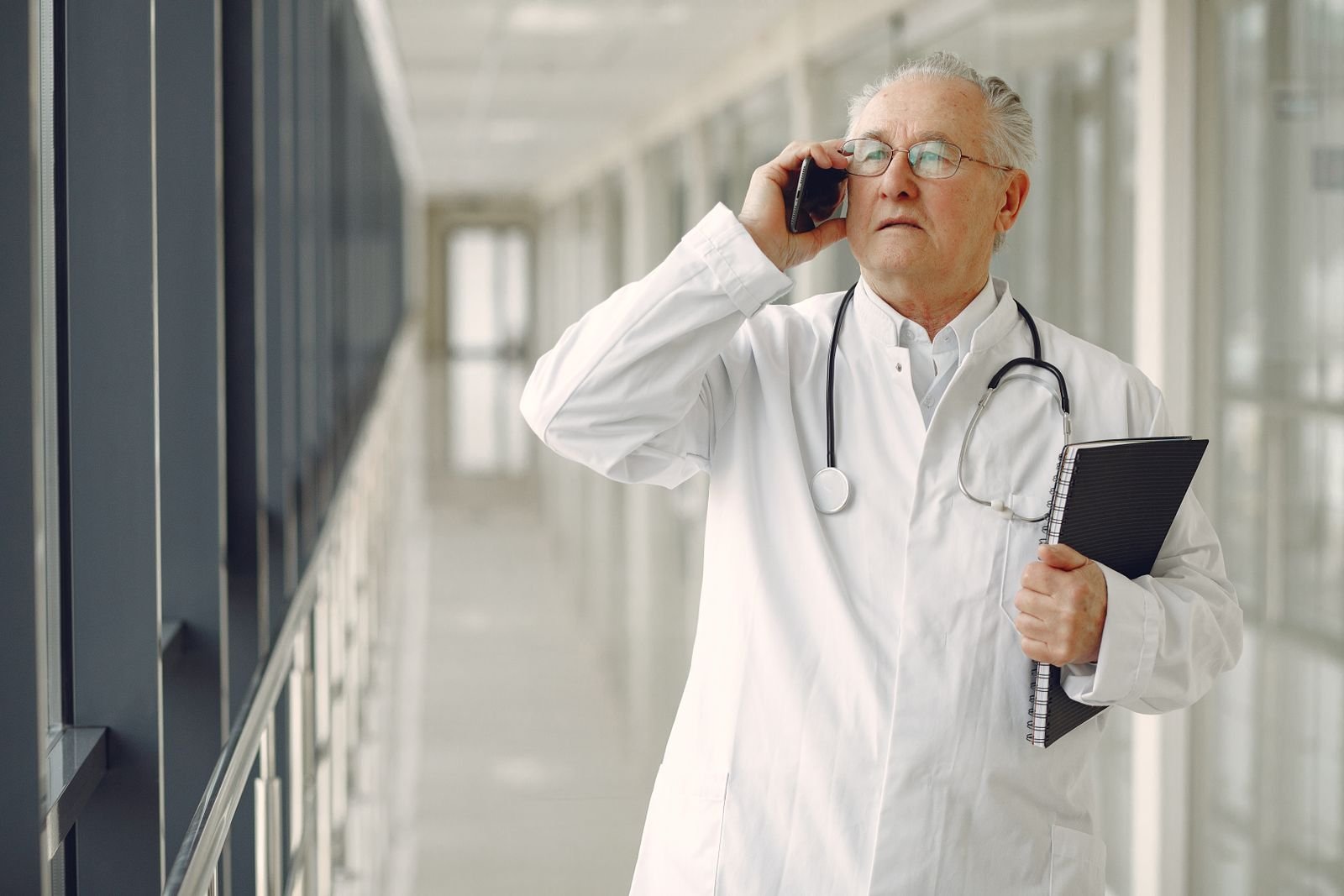 Médico de jaleco branco com estetoscópio no pescoço fala ao celular em corredor de hospital, foto de arquivo