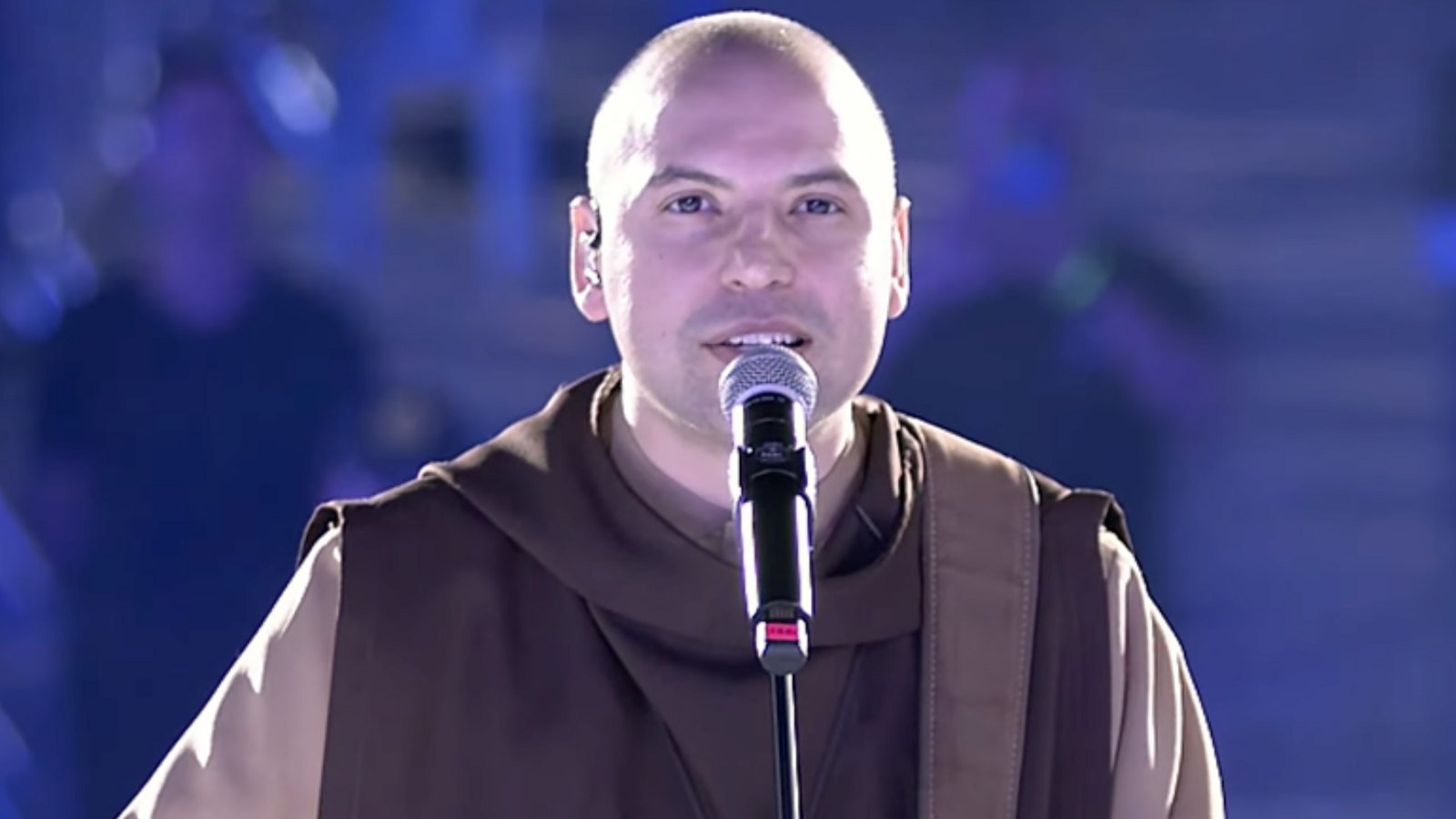 Frei Gilson, de hábito marrom, canta ao microfone com um violão em apresentação