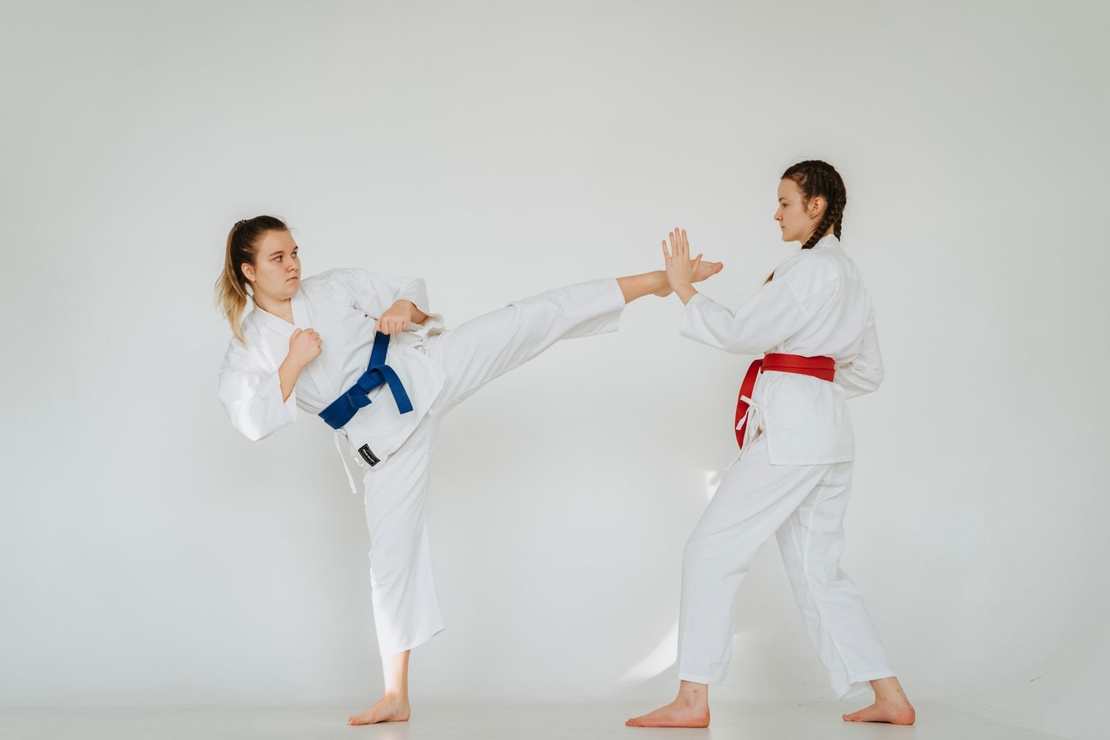Duas lutadoras de taekwondo em quimono branco treinam um chute alto em estúdio com fundo claro (imagem ilustrativa)