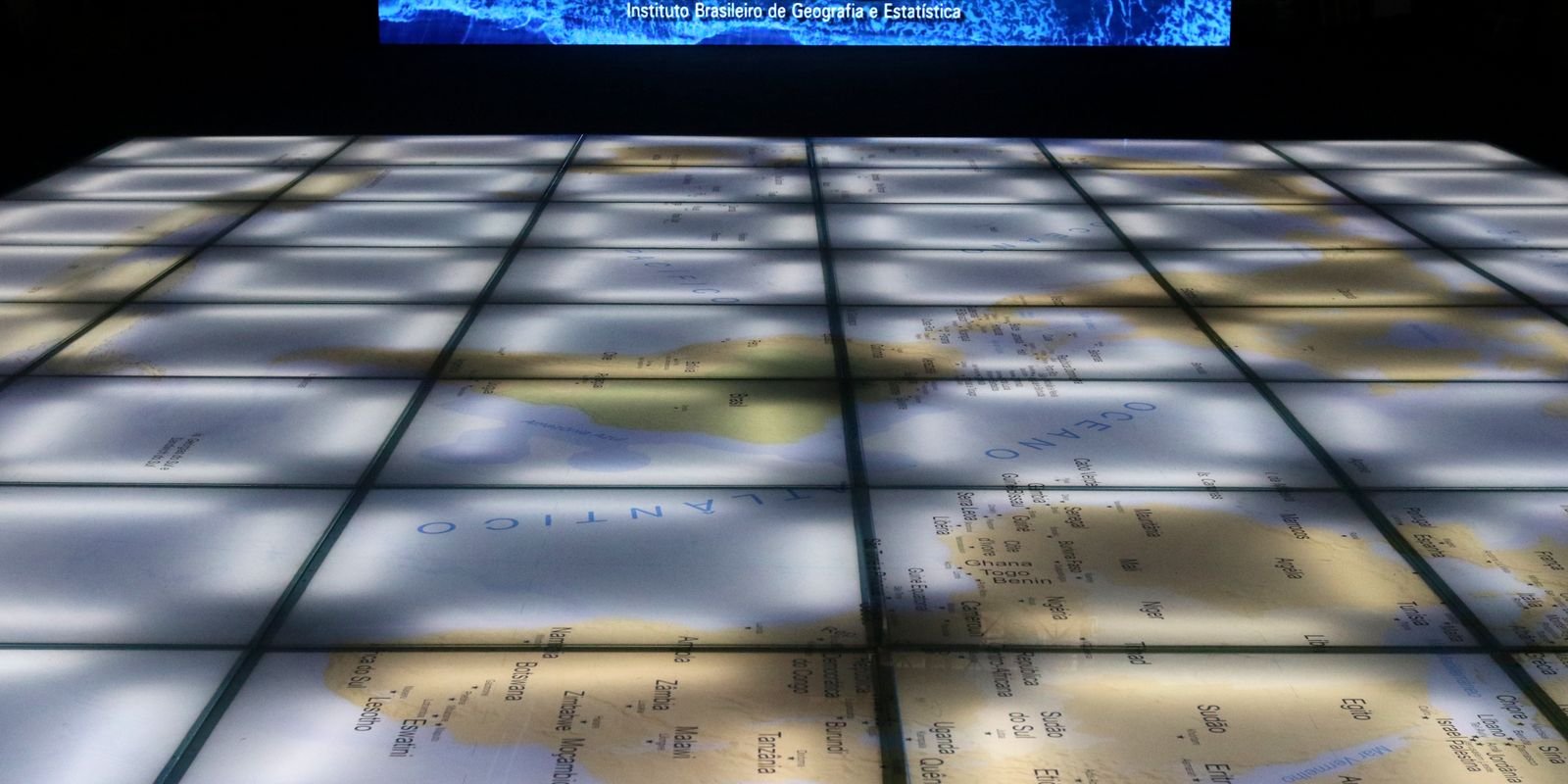 Mapa-múndi iluminado no piso de um espaço expositivo, com o nome do Instituto Brasileiro de Geografia e Estatística ao fundo