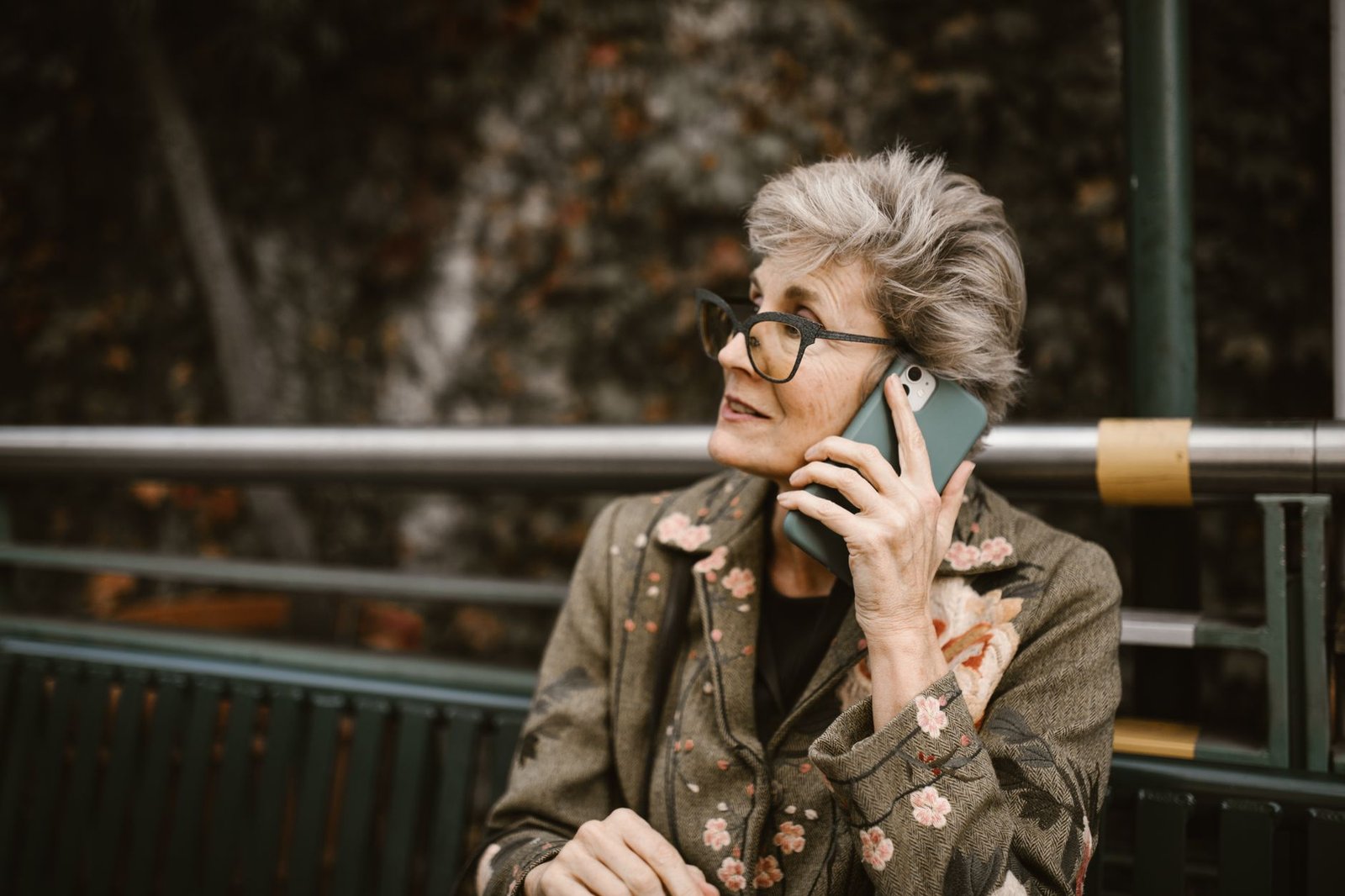 Mulher idosa de óculos conversa ao telefone celular, sentada ao ar livre (imagem ilustrativa)