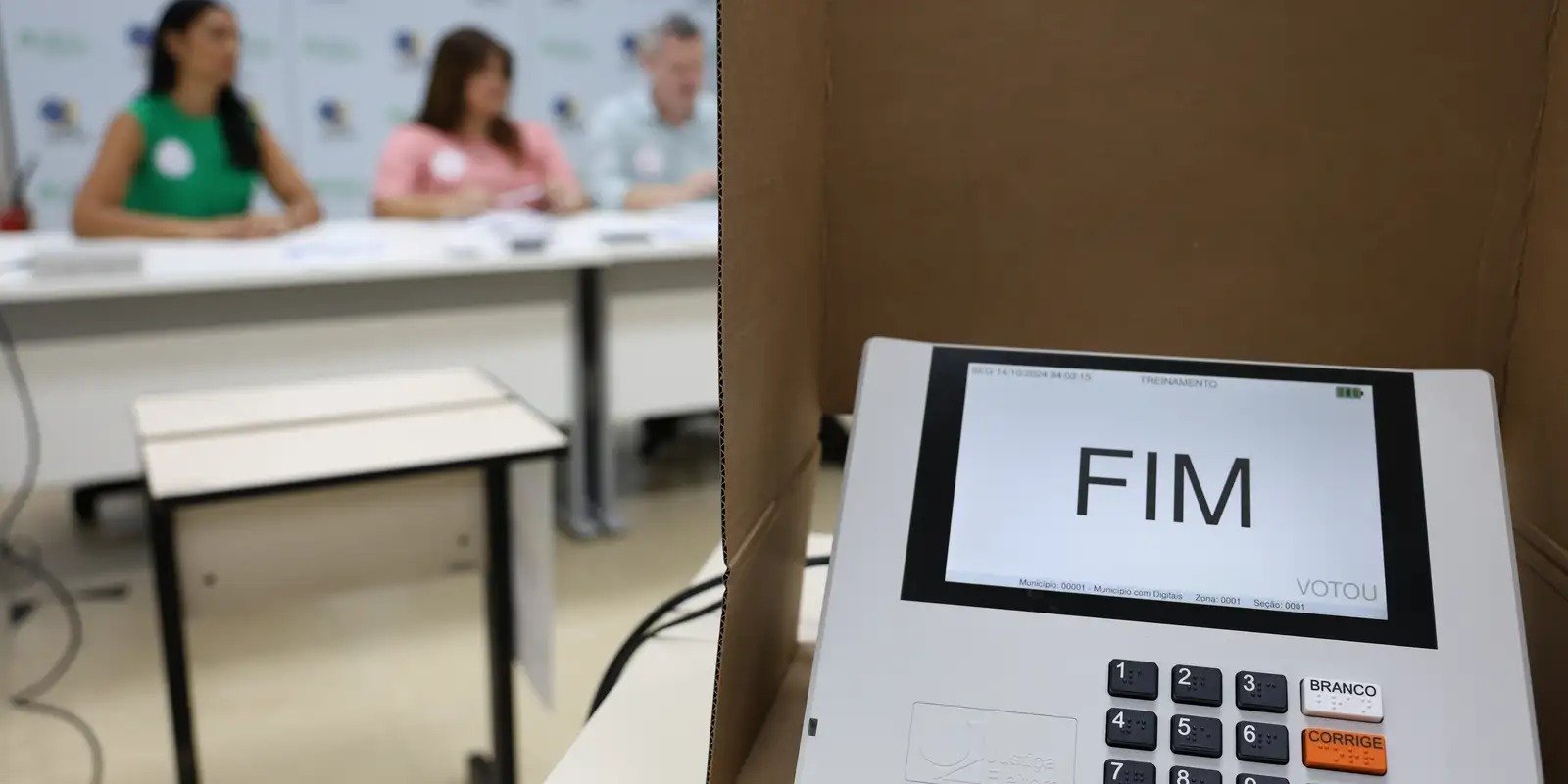 Urna eletrônica com teclas em braile e mesários ao fundo, em seção de treinamento da Justiça Eleitoral